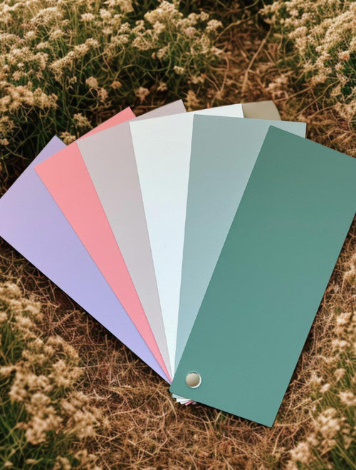 A fanned-out set of six paint color swatches in pastel shades rests on dry grass and surrounded by small white wildflowers. The swatches include lavender, pink, gray, white, blue, and green.