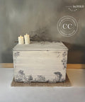 An off-white painted wooden chest with intricate black illustrations from the Cottage Toile Transfer, placed against a gray wall. The chest has two lit small white candles and a small branch on top. A jute mat lies underneath it. A logo for "Dixie Belle Paint Company Content Creator" is seen on the picture.