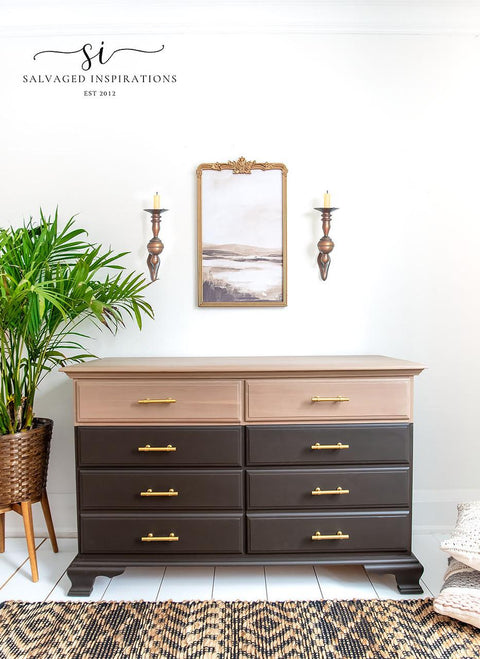 A stylish room featuring a two-tone dresser with brass handles, a woven plant basket on the left with a tall plant, a landscape mirror above the dresser, and two wall-mounted candle holders on either side. A patterned rug lies on the floor.