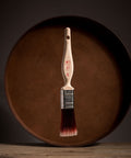 Paint brush with wooden handle and red bristle head floating in front of a dark brown circular platform.