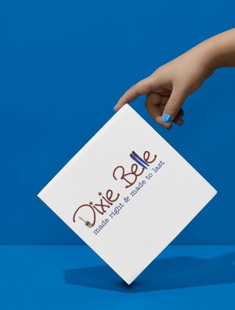 A hand with blue nail polish holds a white square box against a blue background. The box reads "Dixie Belle made right &amp; made to last" in stylized text.