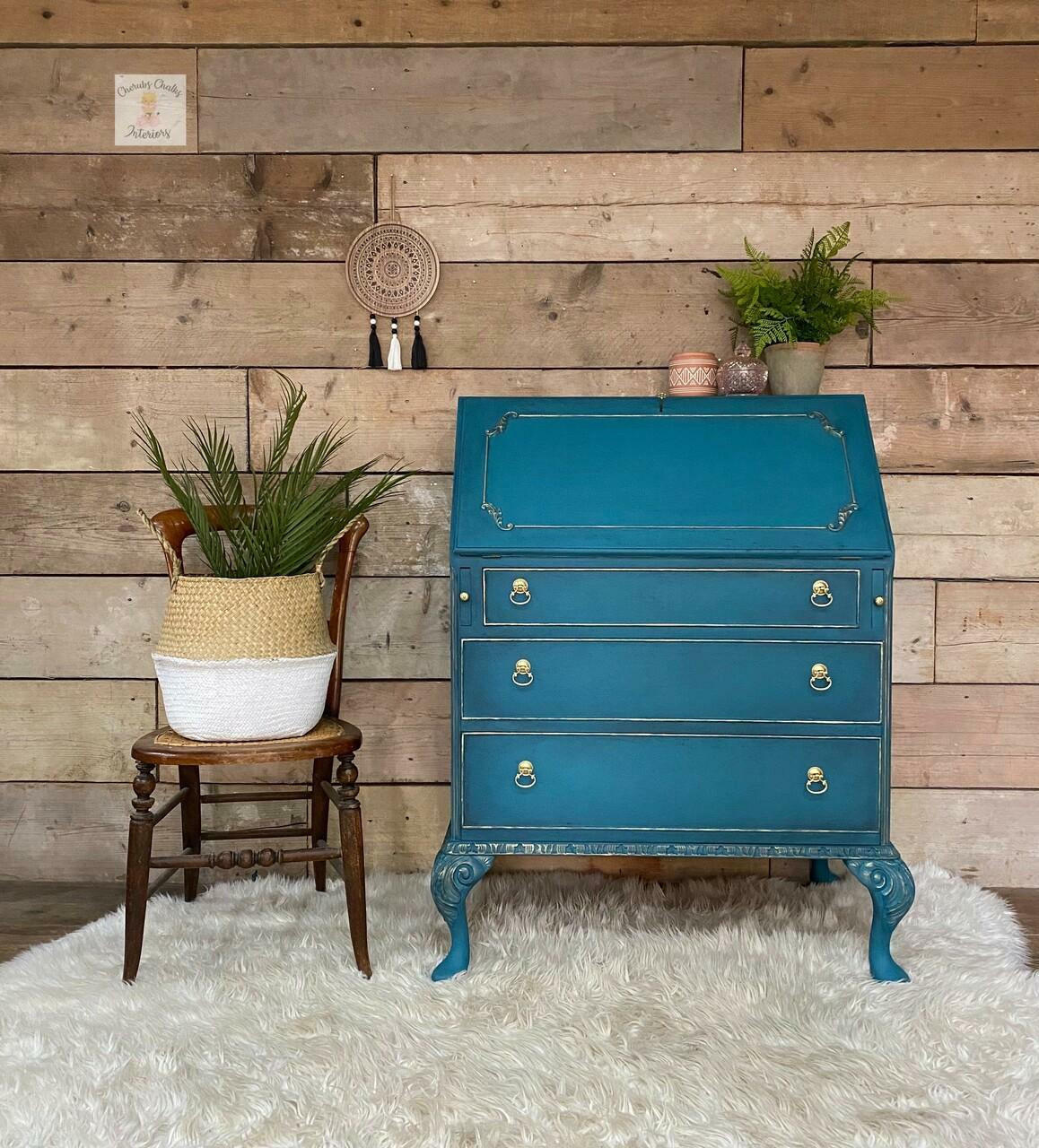 Dixie Belle Paint Antebellum Blue Chalk Mineral Paint is the perfect paint for any DIY project! Classic Drop Lid Desk painted with Antebellum Blue Chalk Mineral Paint. Accented with gold hardware and staged with chair and planter against hardwood wall.