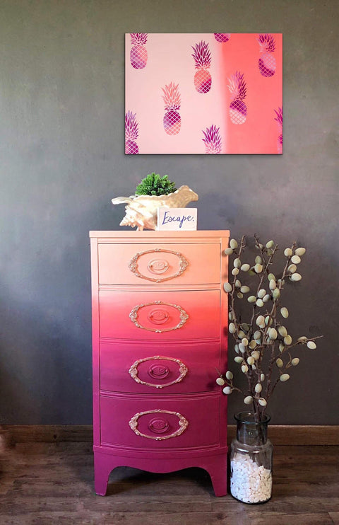 Dixie Belle Paint Apricot Chalk Mineral Paint is the perfect paint for any DIY project!
Chest of drawers painted with Apricot Chalk Mineral Paint and transitions to a purple at the bottom of the chest. Staged  with a seashell and greenery on top with a sign that says " ESCAPE", there's a painting with pineapples on the canvas hanging on the wall with the same colors of the piece.