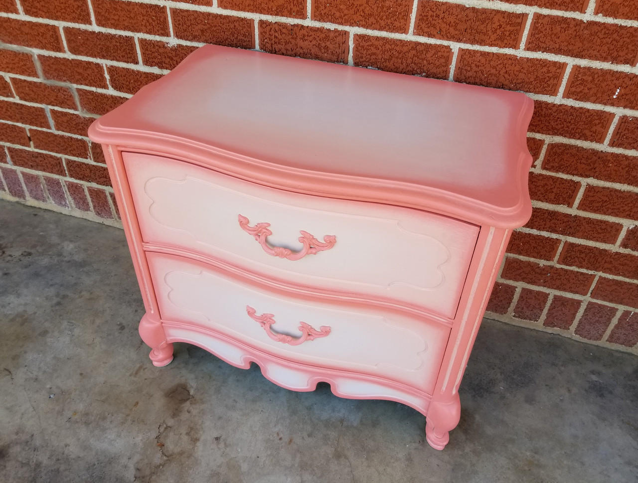 Dixie Belle Paint Apricot Chalk Mineral Paint is the perfect paint for any DIY project!
Bedside table with the body painted in Apricot Chalk Mineral Paint with the top and drawers painted in a blended white. Staged on a brick wall and concrete floor.