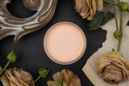 Dixie Belle Paint Apricot Chalk Mineral Paint is the perfect paint for any DIY project!
Open container of Apricot Chalk Mineral Paint staged around the container with ripped pages, tan roses on top of a black background.