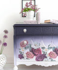 Dixie Belle Paint Aubergine Chalk Mineral Paint is the perfect paint for any DIY project! This image depicts a beautiful armoire painted with Aubergine Chalk Mineral Paint. The piece is multi toned fading from dark to light from the top to the bottom. The piece has been accented with white hardware and our Floral Romance transfer. It is staged with books and a purple flower arrangement.