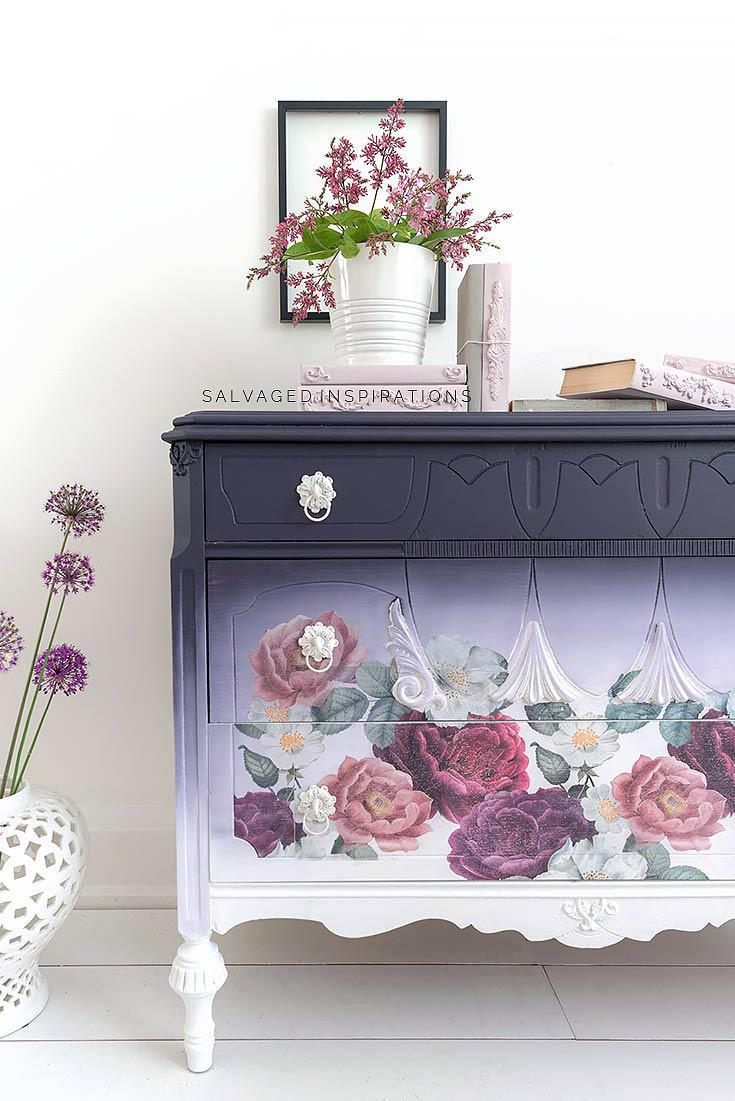 Dixie Belle Paint Aubergine Chalk Mineral Paint is the perfect paint for any DIY project! This image depicts a beautiful armoire painted with Aubergine Chalk Mineral Paint. The piece is multi toned fading from dark to light from the top to the bottom. The piece has been accented with white hardware and our Floral Romance transfer. It is staged with books and a purple flower arrangement.