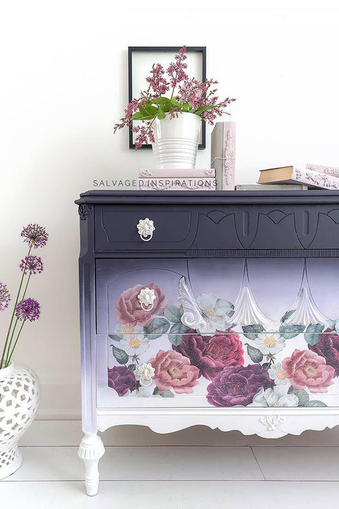 Dixie Belle Paint Aubergine Chalk Mineral Paint is the perfect paint for any DIY project! This image depicts a beautiful armoire painted with Aubergine Chalk Mineral Paint. The piece is multi toned fading from dark to light from the top to the bottom. The piece has been accented with white hardware and our Floral Romance transfer. It is staged with books and a purple flower arrangement.