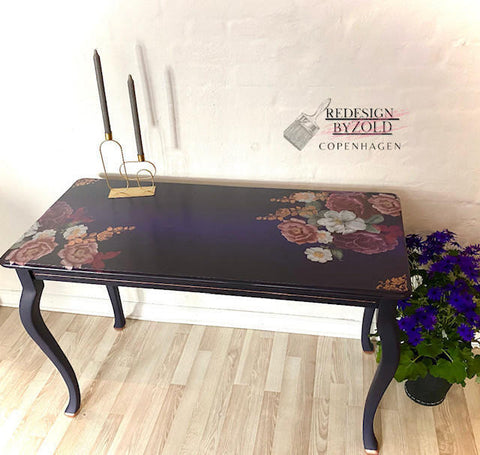 Dixie Belle Paint Aubergine Chalk Mineral Paint is the perfect paint for any DIY project! Aubergine Chalk Mineral Paint on side table accented with our Floral Romance transfer. The piece is staged with a blue and green flower arrangement and candle holders against a white brick wall.