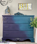 Dixie Belle Paint Aubergine Chalk Mineral Paint is the perfect paint for any DIY project! This tall dresser is painted with Aubergine Chalk Mineral Paint on the left of the dresser and then fades to a light blue. The hardware is painted in the same colors as the cabinet and it is staged with a cork rope and a flower arrangement against a white wall with a picture on it.
