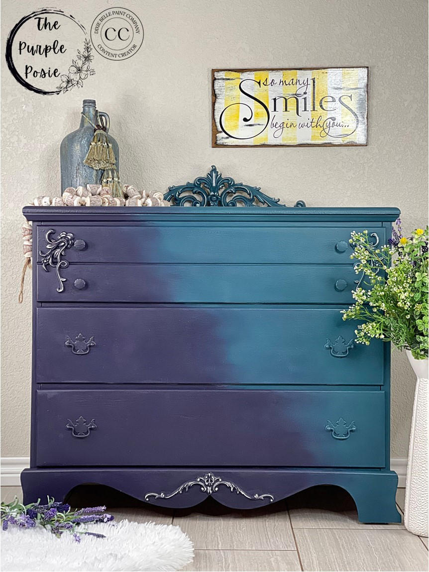 Dixie Belle Paint Aubergine Chalk Mineral Paint is the perfect paint for any DIY project! This tall dresser is painted with Aubergine Chalk Mineral Paint on the left of the dresser and then fades to a light blue. The hardware is painted in the same colors as the cabinet and it is staged with a cork rope and a flower arrangement against a white wall with a picture on it.