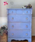 Dixie Belle Paint Blueberry Chalk Mineral Paint is the perfect paint for any DIY project!
 Tall Dresser painted in Blueberry Chalk Mineral Paint staged with purple flowers on top piece. A woven basket with similar flowers besides furniture.