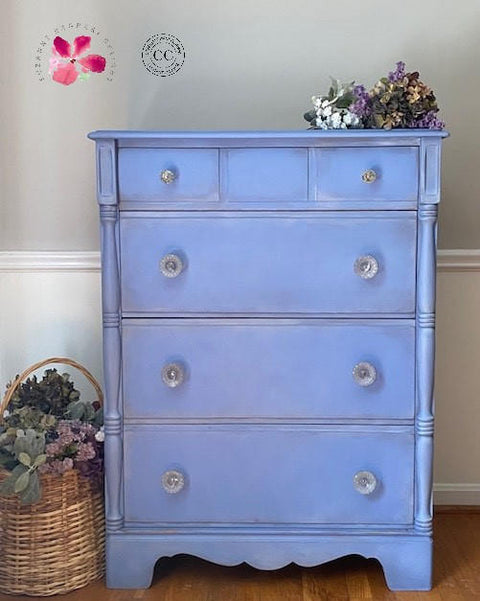 Dixie Belle Paint Blueberry Chalk Mineral Paint is the perfect paint for any DIY project!
 Tall Dresser painted in Blueberry Chalk Mineral Paint staged with purple flowers on top piece. A woven basket with similar flowers besides furniture.