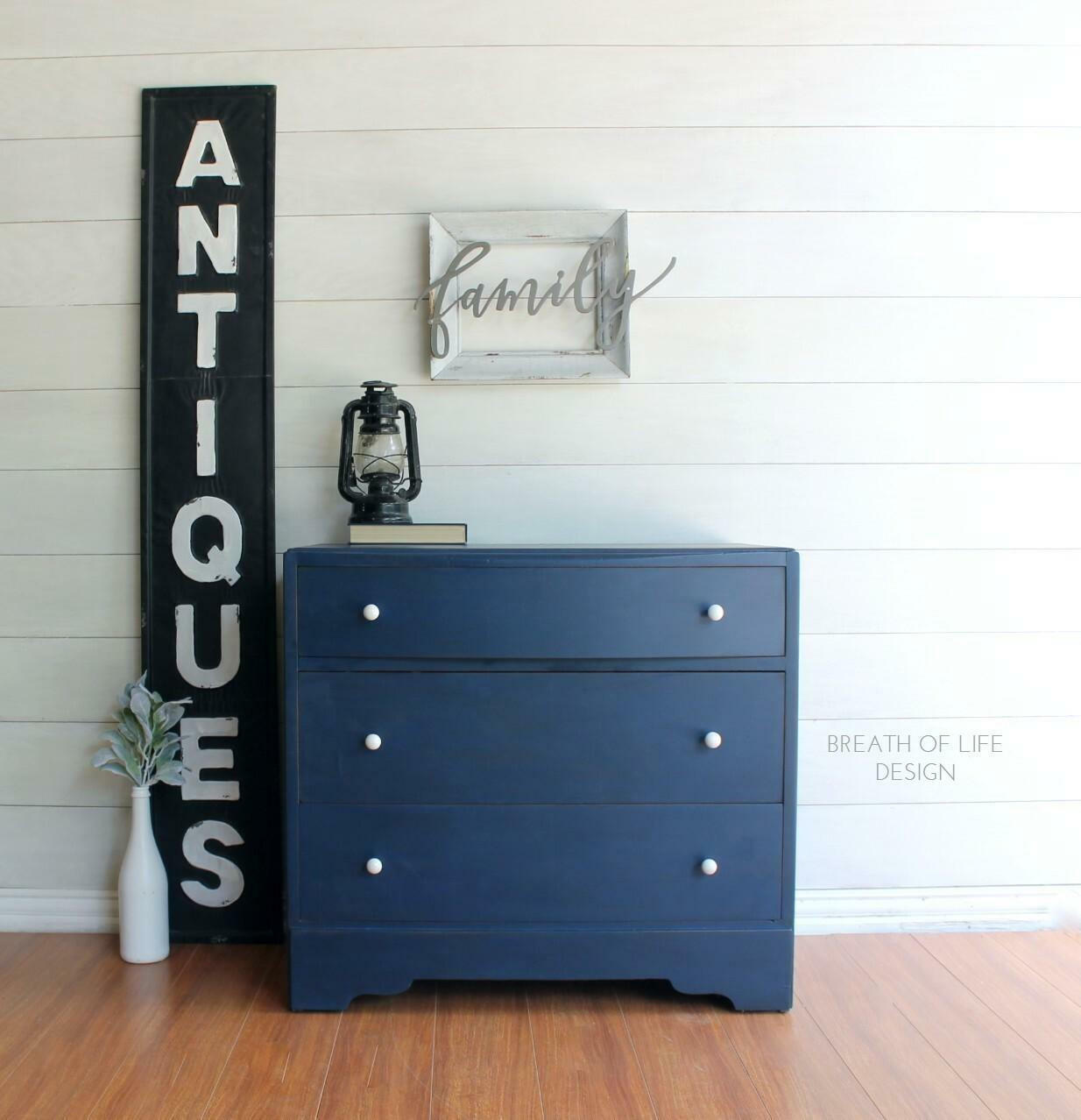 Dixie Belle Paint Bunker Hill Blue Chalk Mineral Paint is the perfect paint for any DIY project!
Dresser painted in Bunker Hill Blue Chalk Mineral Paint with white hardware staged on top of wood floors and walls, Next to piece is a large vertical sign that says "ANTIQUES" and a small white vase with greenery. On top of the piece has a black oil lamp and a book with a white frame that says "FAMILY" hanging above the piece.
