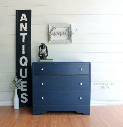 Dixie Belle Paint Bunker Hill Blue Chalk Mineral Paint is the perfect paint for any DIY project!
Dresser painted in Bunker Hill Blue Chalk Mineral Paint with white hardware staged on top of wood floors and walls, Next to piece is a large vertical sign that says "ANTIQUES" and a small white vase with greenery. On top of the piece has a black oil lamp and a book with a white frame that says "FAMILY" hanging above the piece.