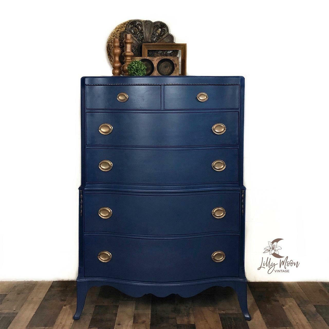 Dixie Belle Paint Bunker Hill Blue Chalk Mineral Paint is the perfect paint for any DIY project!
Chest of drawer painted in Bunker Hill Blue Chalk Mineral Paint with gold hardware and brown wooden decor on top of the piece. Staged in front of a white wall and brown wooden floors.