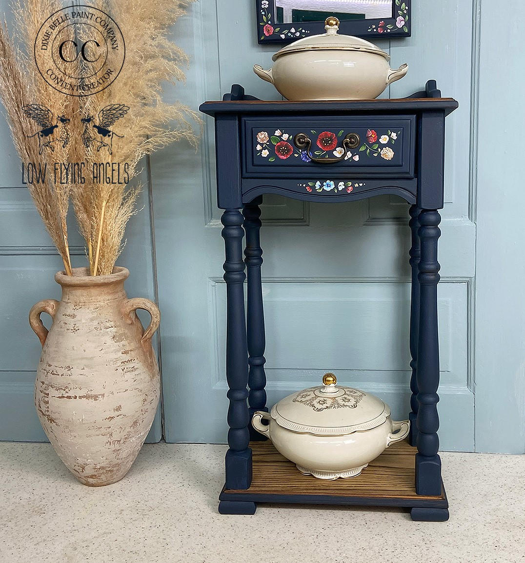 Dixie Belle Paint Bunker Hill Blue Chalk Mineral Paint is the perfect paint for any DIY project!
Beautiful end table painted in Bunker Hill Blue Chalk Mineral Paint with the Dewy Morning Transfer on the front drawer. Staged with two bowls on the top and the bottom shelf with a large tan ceramic vase with large branches with feathers on it to the left side of the piece.