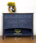 Dixie Belle Paint Bunker Hill Blue Chalk Mineral Paint is the perfect paint for any DIY project!
Credenza painted in Bunker Hill Blue Chalk Mineral Paint with a dry brush distressed look with neutral hardware. Staged with sunflowers inside a metal vase on top of piece and a woven bowl full of greenery at the bottom shelf.