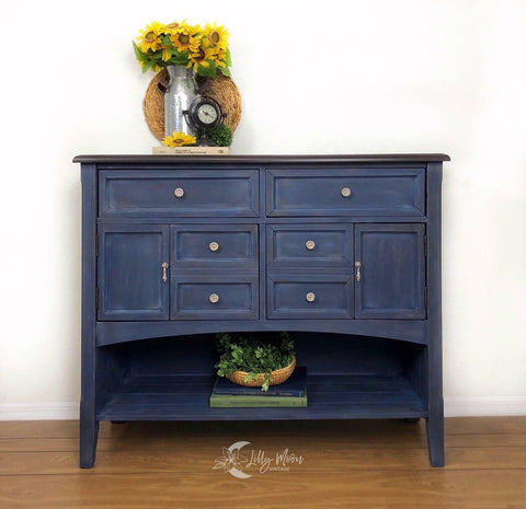 Dixie Belle Paint Bunker Hill Blue Chalk Mineral Paint is the perfect paint for any DIY project!
Credenza painted in Bunker Hill Blue Chalk Mineral Paint with a dry brush distressed look with neutral hardware. Staged with sunflowers inside a metal vase on top of piece and a woven bowl full of greenery at the bottom shelf.