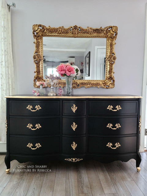 Dixie Belle Paint Caviar Chalk Mineral Paint is the perfect paint for any DIY project!
Dresser painted in Caviar Chalk Mineral Paint with a natural wood colored top and gold hardware. Staged with glass containers and vase with pink flowers and a large gold framed mirror hanging above the piece.