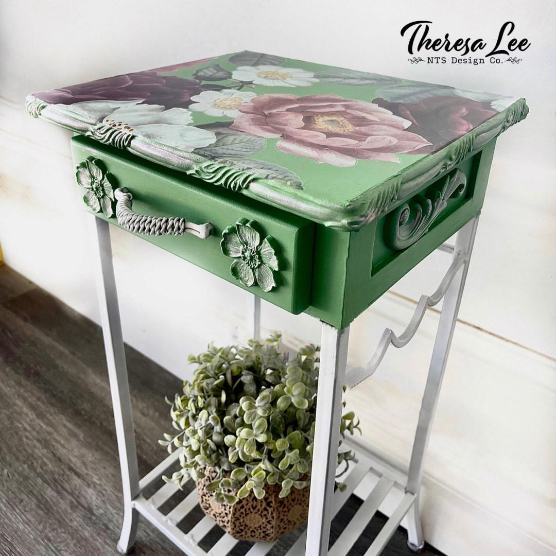 Dixie Belle Paint Chameleon Wax gives your furniture DIY project an iridescent glow and is sheer wax utilized to unveil the  highlight of your project!
Bedside table painted in green and white legs with Chameleon Wax along the edge and applique . As well as the Floral Romance Transfer on the top of table. Staged with a potted faux succulent under the table.