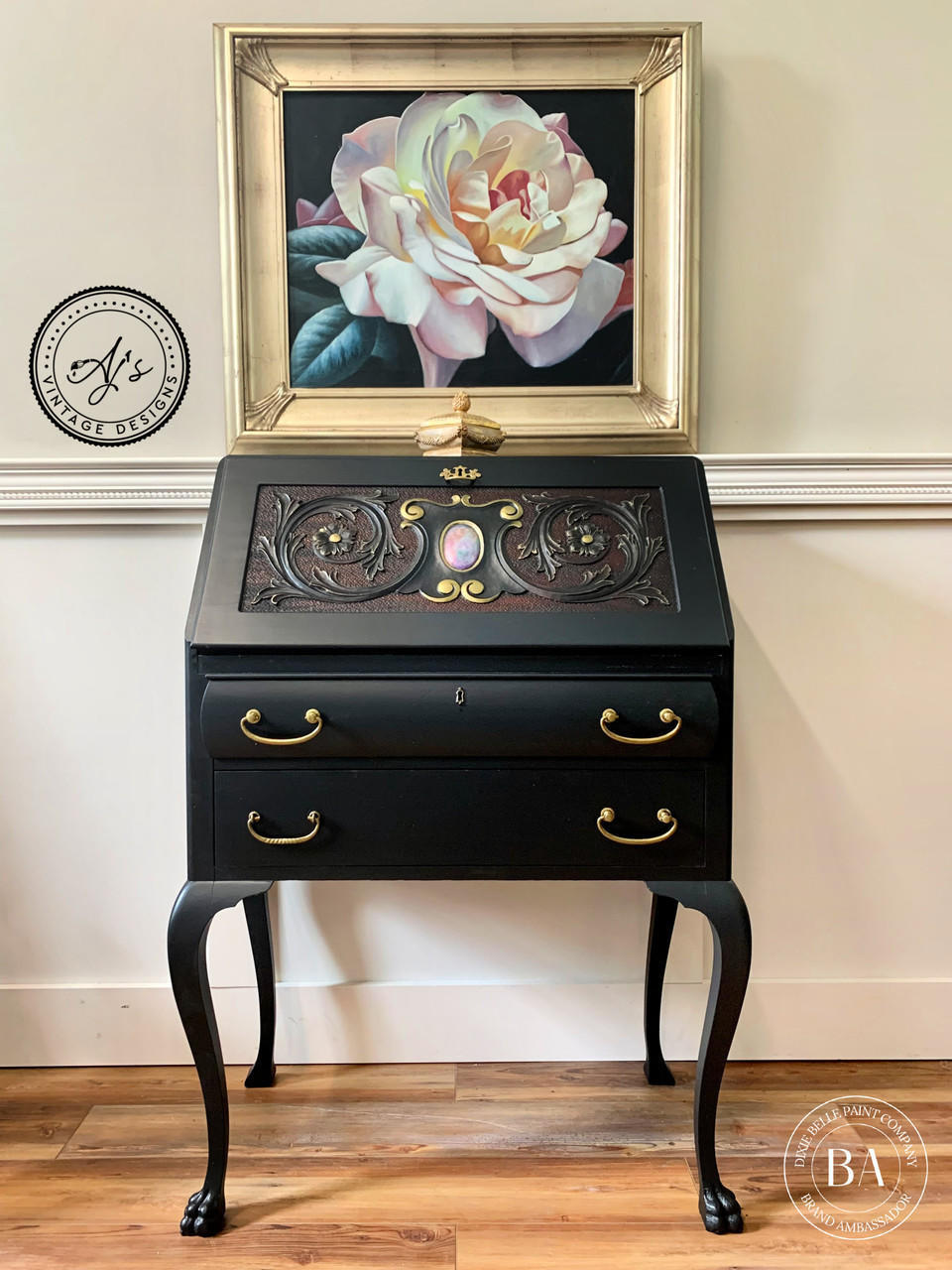 Dixie Belle Paint Chameleon Wax gives your furniture DIY project an iridescent glow and is sheer wax utilized to unveil the  highlight of your project!
Sleek secretary desk with the  Chameleon Wax on the desk panel. It has gold hardware with an opal centerpiece. Staged with a gold picture from of a pink flower is hanging above piece.