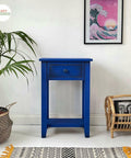 Dixie Belle Paint Cobalt Blue Chalk Mineral Paint is the perfect paint for any DIY project!
There is a small table at bar stool height painted completely in Cobalt Blue CMP next to a white wall.  There are two plants to the left of the table and slightly in front of the table.  One is a palm and the other a cactus in planters.  To  the right of the table is wicker magazine holder with a magazine in it with a picture of a man's face.  On the wall is a pink painting of a large wav and the sun going down.