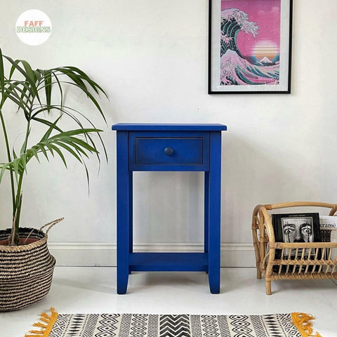 Dixie Belle Paint Cobalt Blue Chalk Mineral Paint is the perfect paint for any DIY project!
There is a small table at bar stool height painted completely in Cobalt Blue CMP next to a white wall.  There are two plants to the left of the table and slightly in front of the table.  One is a palm and the other a cactus in planters.  To  the right of the table is wicker magazine holder with a magazine in it with a picture of a man's face.  On the wall is a pink painting of a large wav and the sun going down.