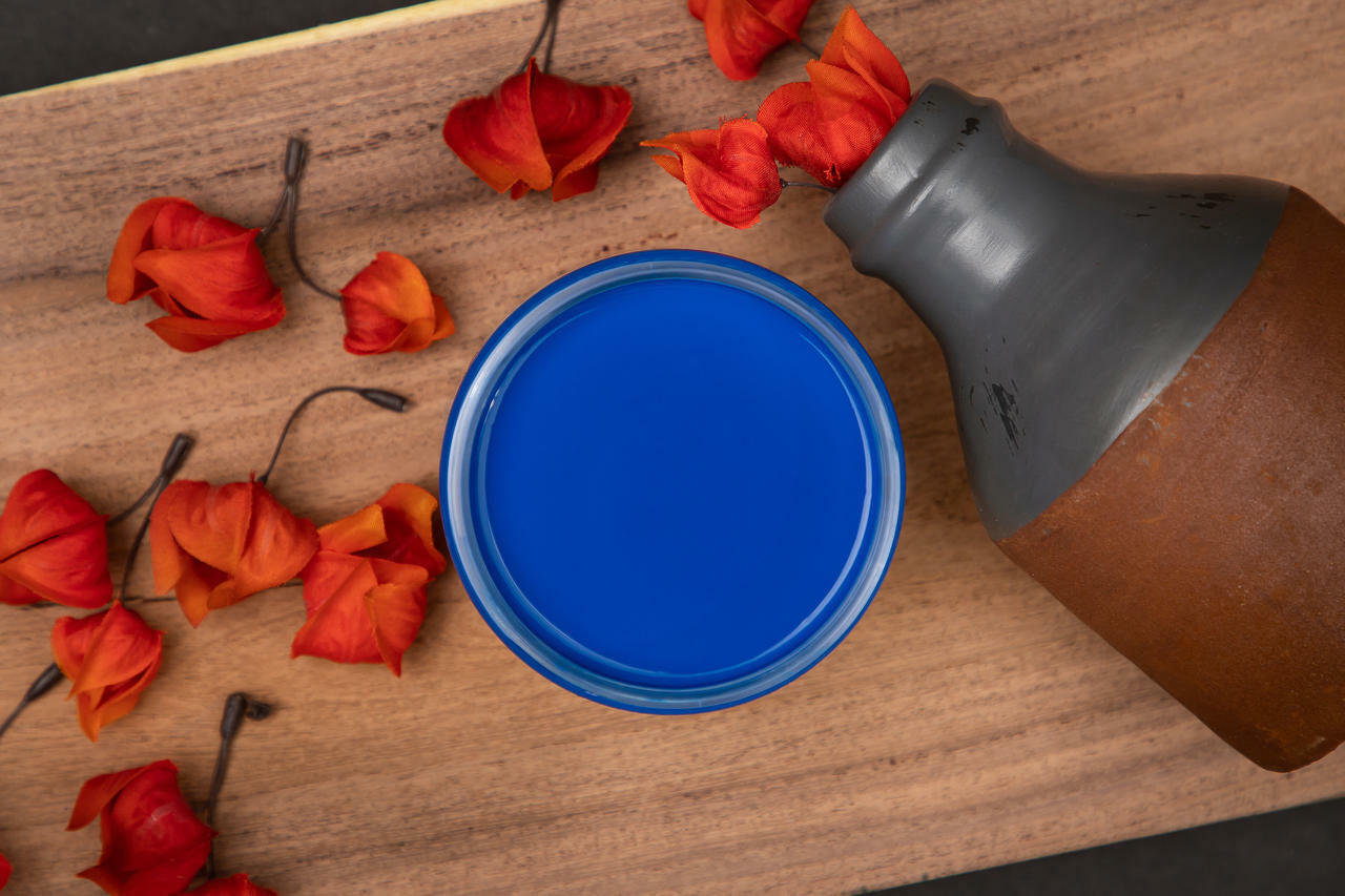 Dixie Belle Paint Cobalt Blue Chalk Mineral Paint is the perfect paint for any DIY project! An open jar of Cobalt Blue Chalk Mineral Paint is placed on a piece of No Pain Gel Stain with orange flowers placed around it and an old rusty canteen placed on it's side.