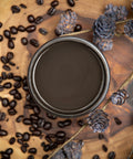 Dixie Belle Paint Coffee Bean Chalk Mineral Paint is the perfect paint for any DIY project!  
There is an open paint can that you are looking down on so you only see the brown paint inside.  The paint can is sitting on a wood table that has natural sides and is stained in natural and dark wood.  There are coffee beans scattered around the paint can.  There dried, tan twigs on the table with dried pine cones scattered around and on the twigs.