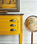 Dixie Belle Paint Colonel Mustard Chalk Mineral Paint is the perfect paint for any DIY project!
This large buffet has metal pulls, wood-u-bend appliques and a dark wood top.  The buffet is painted completely in Colonel Mustard CMP.  To the right of the buffet is a stand alone globe.  on top of the buffet sits two, old books and a leafy plant in a black container.  Above the buffet on the white, ship lap wall is a painting of golden grass with a wooden fence and a few sparse plants next to the fence.