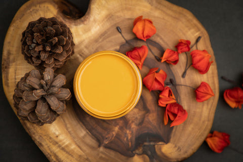 Dixie Belle Paint Colonel Mustard Chalk Mineral Paint is the perfect paint for any DIY project!
You are looking down on a can of Colonel Mustard so you can only see the paint inside.  The paint can is sitting on top of a large wood log.  On top of the log, next to the Colonel Mustard CMP are two pine cones to the left and to the right are orange flower pedals,  two has fallen to to the dark, wood floor.