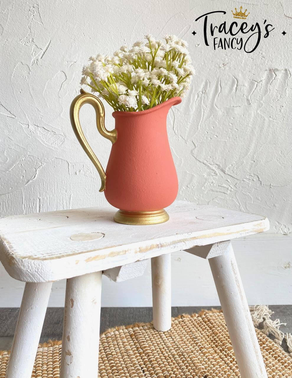Coral chalk painted pitcher with a gold base and handle. Staged with white flowers inside, on a white painted distressed stool.