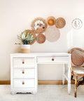 Dixie Belle Paint Cotton Chalk Mineral Paint is the perfect paint for any DIY project!
This is a common desk with a long drawer where you can put a chair and 3 drawers on the left.  This is all painted in Cotton CMP and all the knobs are brown.  There's a plant on top of desk in a white and brown pot.  To the left is a dark, wicker chair with a dark wicker back.  There are six, different sized, wicker, round wall decorations on the wall behind the desk.  The desk is on a white, bushy carpet.