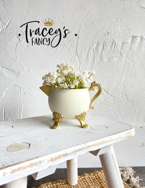 White chalk painted pitcher with gold legs, handle and spout. Staged with white flowers inside, on a white painted distressed stool.