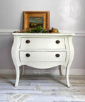 White chalk painted dresser with black handles. Staged with greenery and a gold picture frame on top and book pages strewn on the floor.