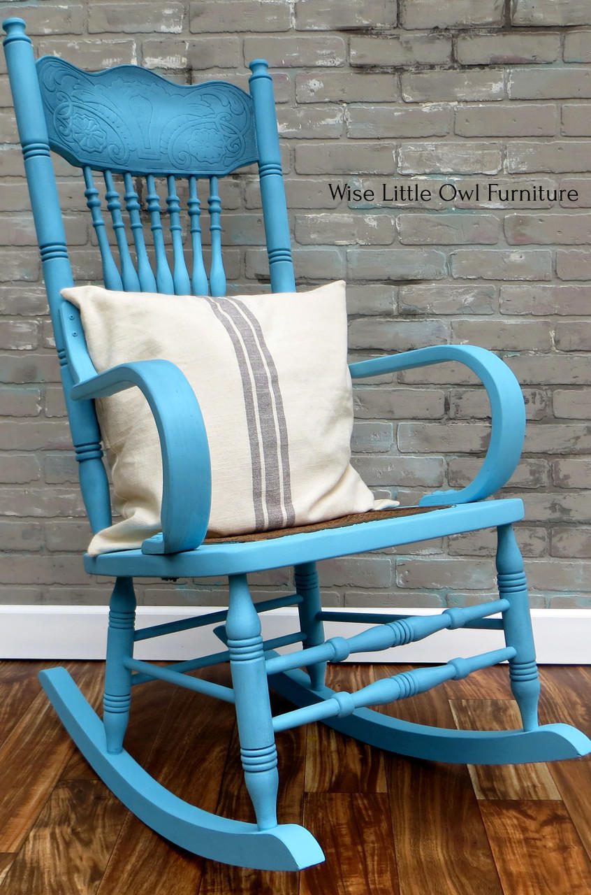 Dixie Belle Paint Dixie Belle Blue Chalk Mineral Paint is the perfect paint for any DIY project!
This is an old fashioned rocking chair painted in Dixie Belle Blue CMP.  There is a cream pillow on the seat of the rocker and the pillow has 3 stipes of brown color.  The chair is in front of a brick wall and rests on a glossy, wood floor.