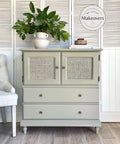 Dixie Belle Paint Dried Sage Chalk Mineral Paint is the perfect paint for any DIY project!
Cabinet with cane webbing doors above drawers painted in Dried Sage Chalk Mineral Paint. Staged on wood floors and a white door panel background. On top has a white vase with handles and greenery.