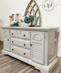 Dixie Belle Paint Driftwood Chalk Mineral Paint is the perfect paint for any DIY project!
Gray chalk painted dresser with black knobs. Three candle sticks, large white vase with flowers, and a wooden distressed arch.