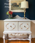 Dixie Belle Paint Driftwood Chalk Mineral Paint is the perfect paint for any DIY project!
Gray chalk painted furniture with a glass vase containing red and green flowers and a vintage perfume bottle. Gold metal lamp with white lamp shade.