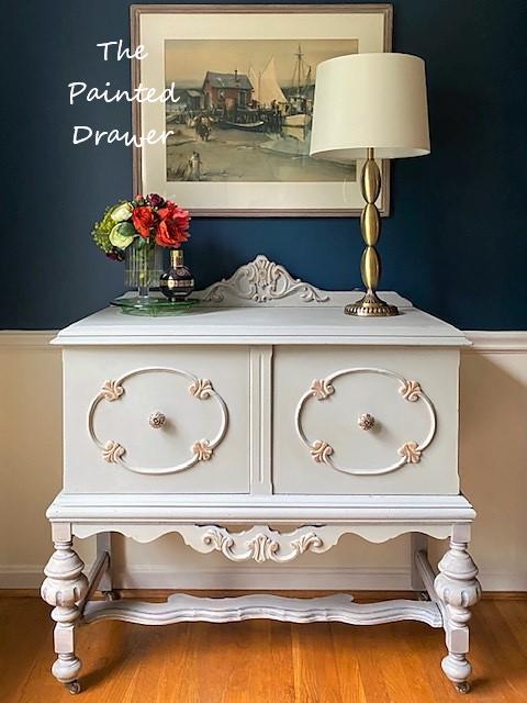 Dixie Belle Paint Driftwood Chalk Mineral Paint is the perfect paint for any DIY project!
Gray chalk painted furniture with a glass vase containing red and green flowers and a vintage perfume bottle. Gold metal lamp with white lamp shade.