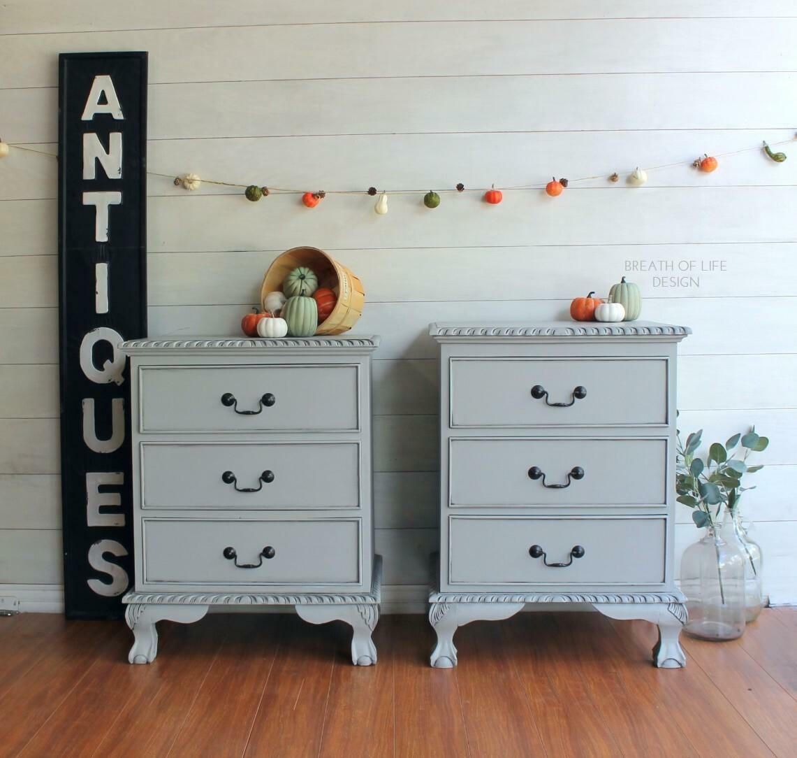 Dixie Belle Paint Driftwood Chalk Mineral Paint is the perfect paint for any DIY project!
A pair of gray chalk painted nightstands staged with fall décor. A large slim black sign with "antiques" written in white letters, faux pumpkins and squash strown on a line above the nightstands, a basket full of faux pumpkins and squash overflowing onto the top of one of the nightstands, and a vase full of faux eucalyptus.