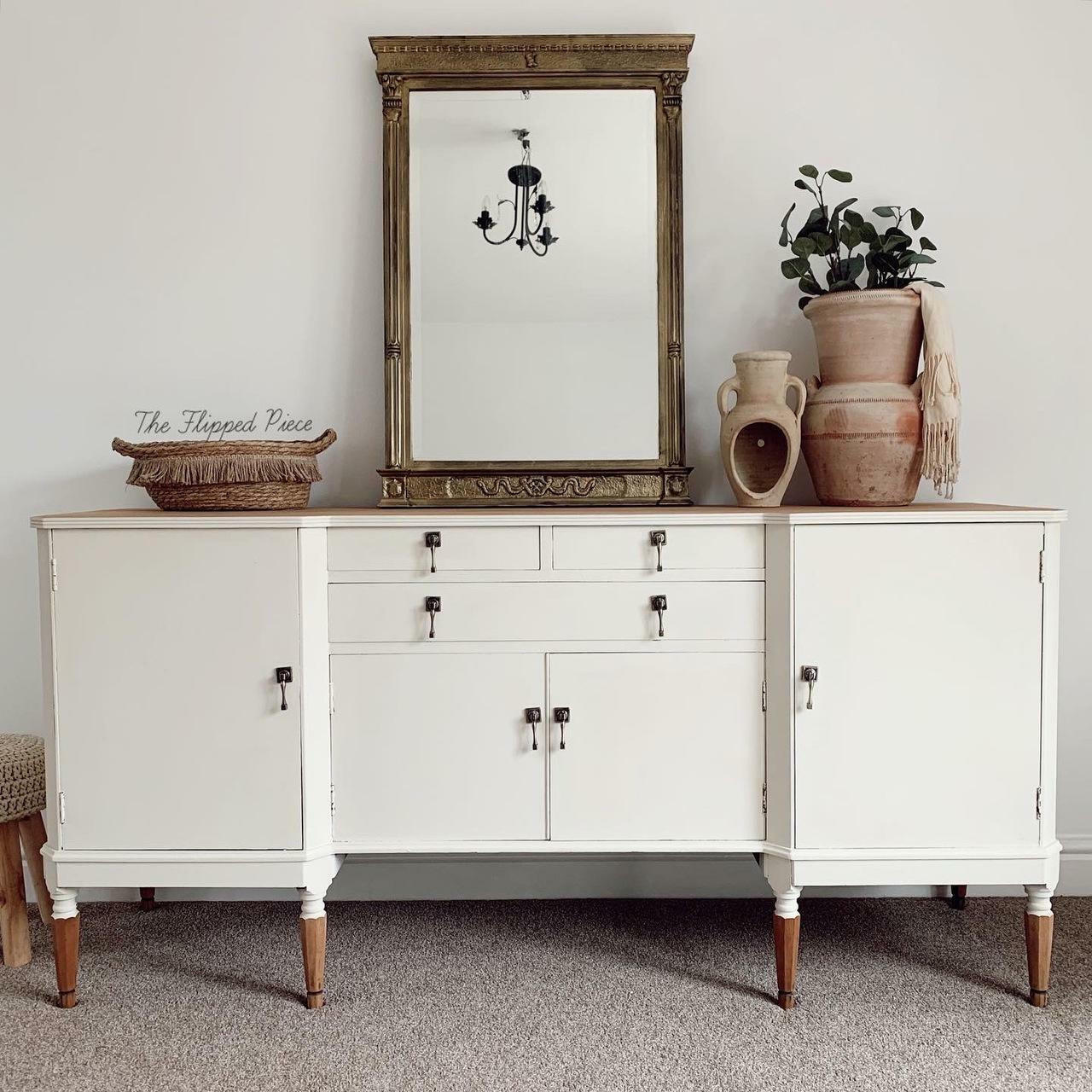 Dixie Belle Paint Drop Cloth Chalk Mineral Paint is the perfect paint for any DIY project! White chalk painted dresser with wooden legs. Staged with a boho style basket, large mirror and 2 ceramic pots, with greenery and a white blanket hanging out of the larger pot.