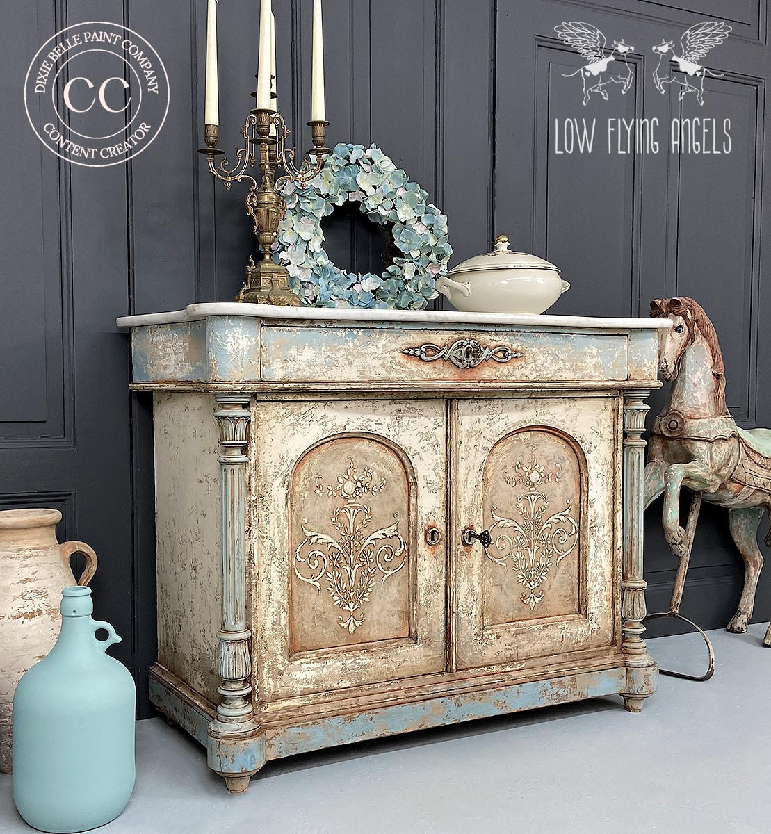 Dixie Belle Paint Dusty Blue Chalk Mineral Paint is the perfect paint for any DIY project! White and blue rustic dresser makeover with large candlesticks, blue flowery wreath, and a large white pot on top. Staged with a large white and blue jug, also a vintage merry-go-round carnival horse.