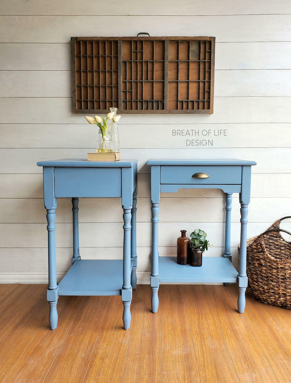 Dixie Belle Paint Dusty Blue Chalk Mineral Paint is the perfect paint for any DIY project! Two blue chalk painted end tables staged with a vase with white flowers set on a book. With a large wooden frame on a white pallet wall. Small glass vases containing greenery and a large woven basket.