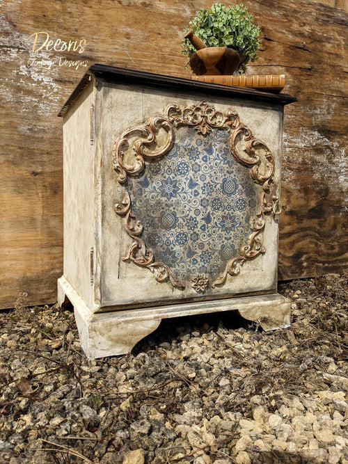 Dixie Belle Paint Easy Peasy™ Spray Wax 8oz. White grungy chalk painted cabinet with a Blue Glass Ornate Rice Decoupage Paper applied on the front center. Dark wooden stained top. Staged with a book on the top with wooden bowls and greenery.