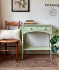 Dixie Belle Paint Farmhouse Green Chalk Mineral Paint is the perfect paint for any DIY project!
Console table painted in Farmhouse Green Chalk Mineral Paint with white hardware, Staged with a wooden chair and a pillow with fringe on it, and books are staged on top of the piece.
