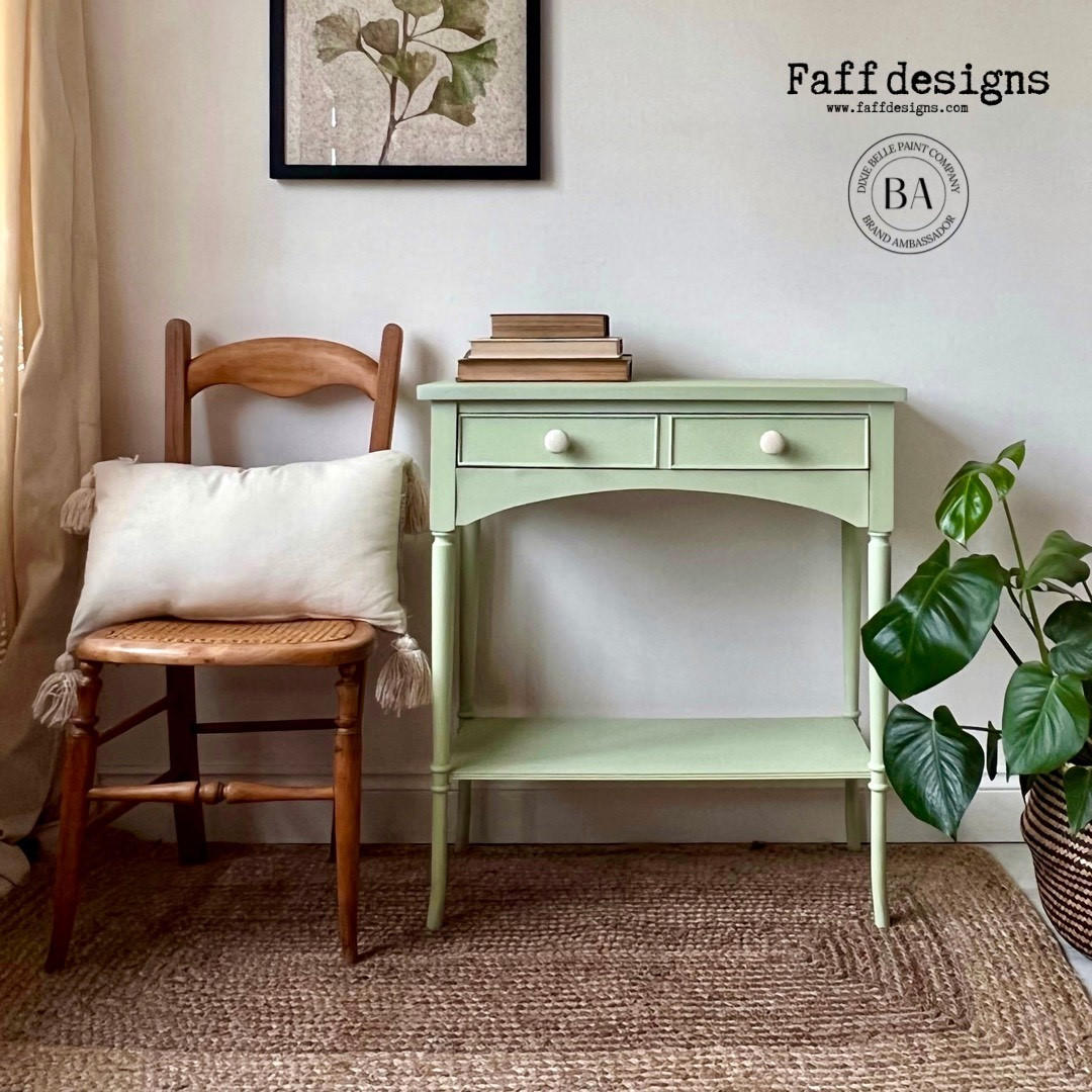 Dixie Belle Paint Farmhouse Green Chalk Mineral Paint is the perfect paint for any DIY project!
Console table painted in Farmhouse Green Chalk Mineral Paint with white hardware, Staged with a wooden chair and a pillow with fringe on it, and books are staged on top of the piece.