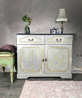 Dixie Belle Paint Gold Shimmer Glaze. White Gray Vanity with Gold Shimmer Glaze used in the indexes of the drawers and doors , to give it an elegant and royal look. Staged with a gold lamp and a porcelain teapot with teacups. Glass see through vase with flowers.