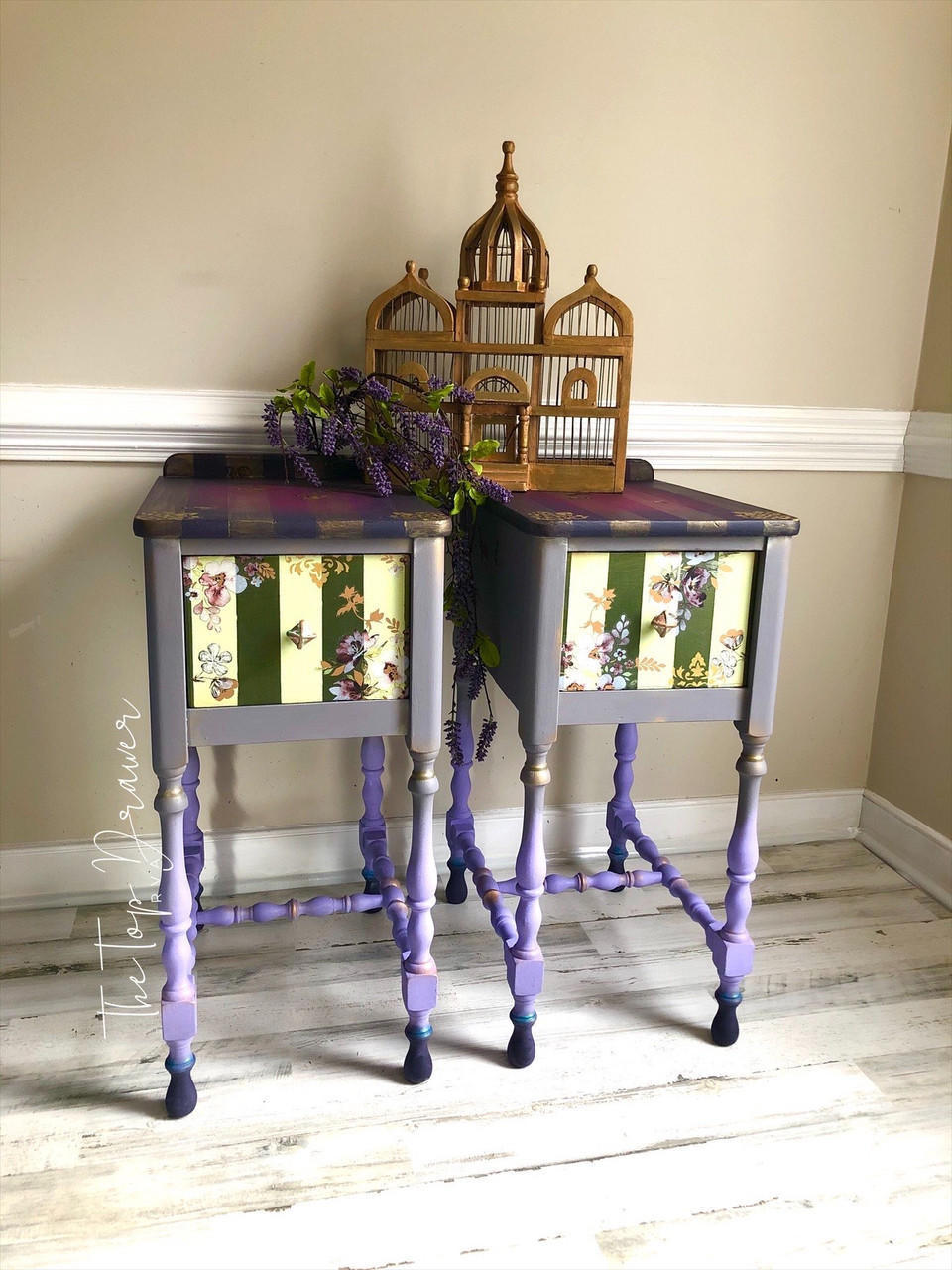 Dixie Belle Paint Gold Shimmer Glaze. Two purple Side tables with Gold shimmer glaze being used to create stripes on top of pieces with gold accents in the nook's of the legs to create more dimension. White and green stripes drawer has floras design around it. Staged on a white washed wood floor with gold temple cage figure on top of tables with a lavender branch hanging in-between tables.